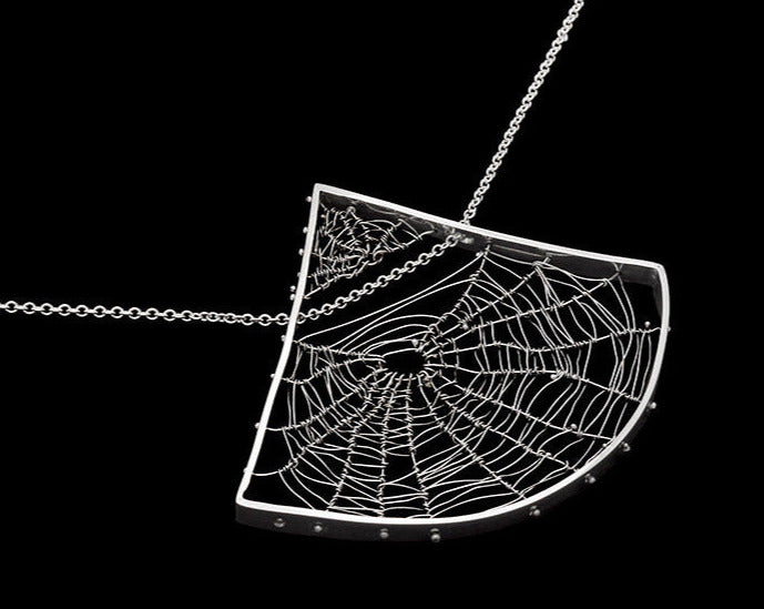 axe-shaped orbweaver spiderweb necklace, hand woven and twined by Peggy Skemp. The necklace was made in solid silver, rhodium plated. The photo has a black background and was taken by Chicago photographer, Rachel Hanel.